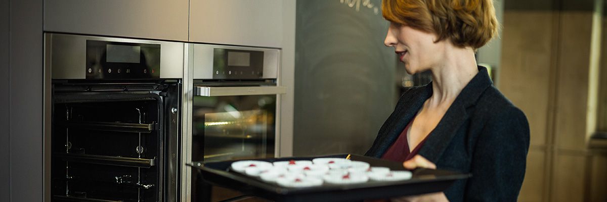 Eine Frau hält ein Backblech mit kleinen Förmchen und schiebt es in einen offenen Neff Backofen in moderner Küche.